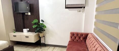 En moderne stue med en beige sofa, en brun lænestol, et fladskærms-tv monteret på en mørk trævæg, en aircondition og en potteplante.