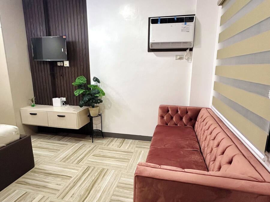 A modern living room with a beige sofa, a brown couch, a flat-screen TV mounted on a dark wooden wall, an air conditioning unit, and a potted plant.