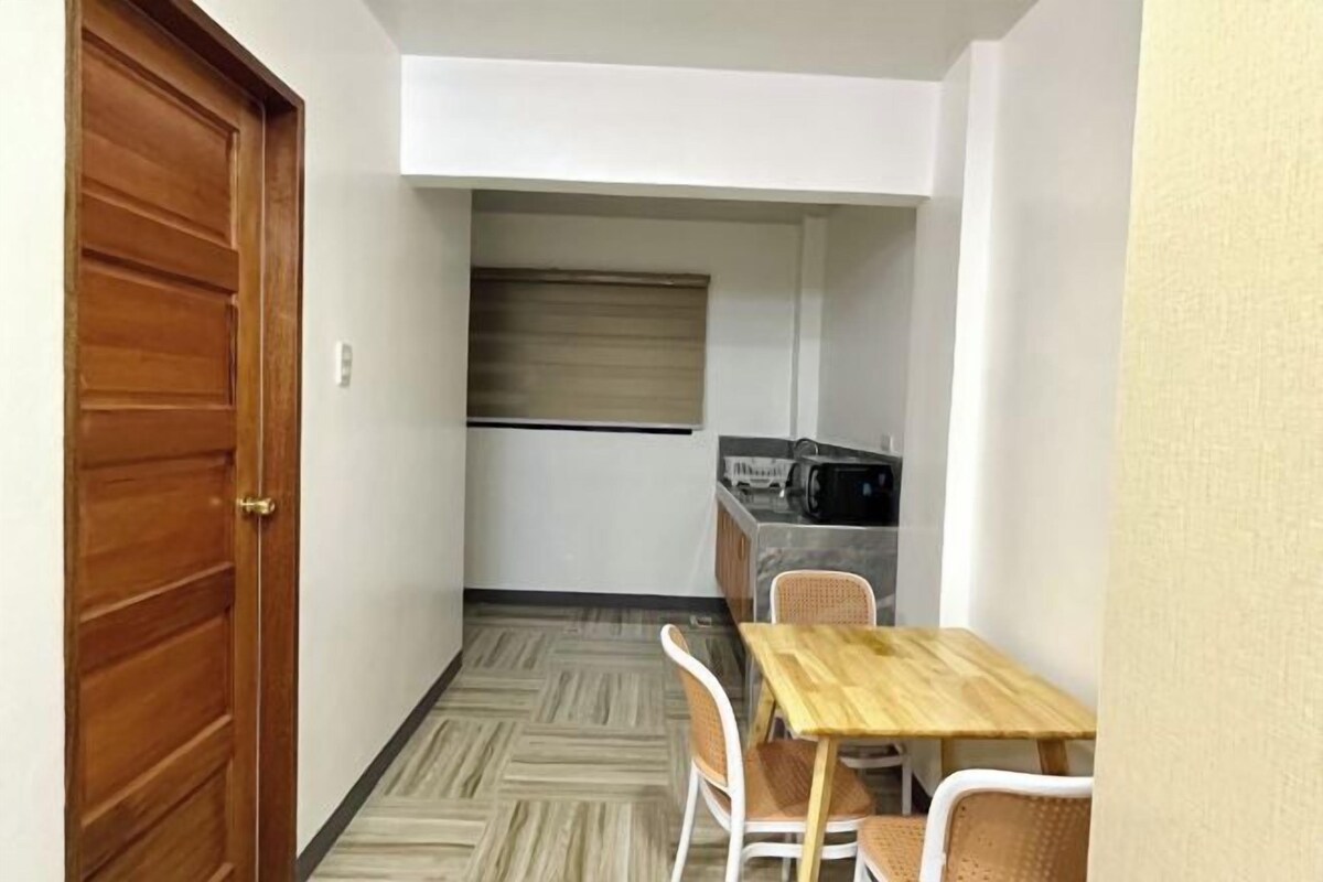 A compact room with a wooden door, a small kitchenette, a dining table with chairs, and a patterned floor.
