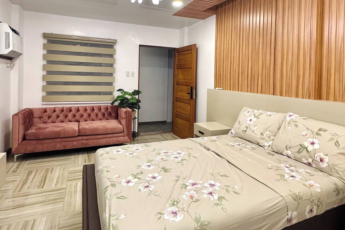 A bedroom with a bed, a sofa, a wooden panel wall, a ceiling light, and a door.