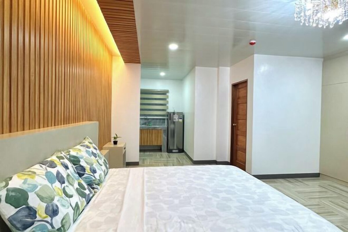 A hotel room with a bed, wooden panel wall, a chandelier, a kitchenette, and a door.