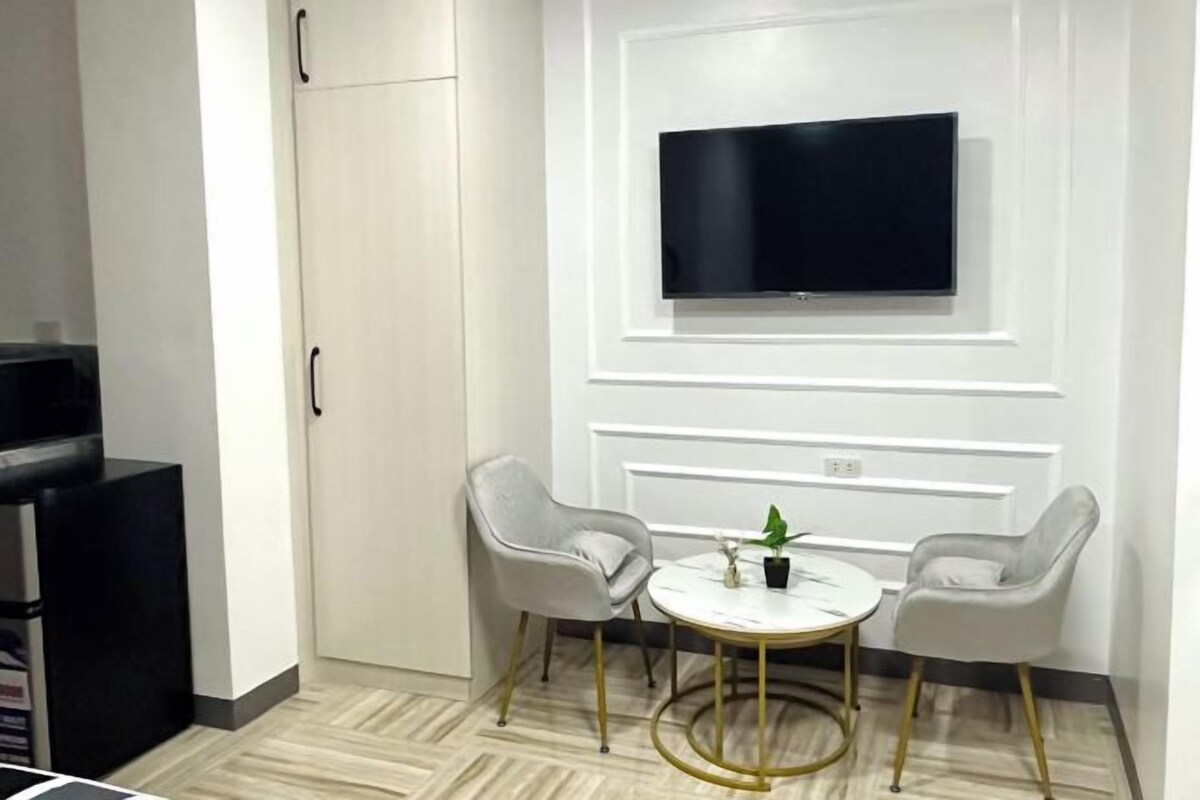 A modern room with a TV mounted on a white wall, two grey chairs around a small round table with a plant, and a black cabinet to the left.