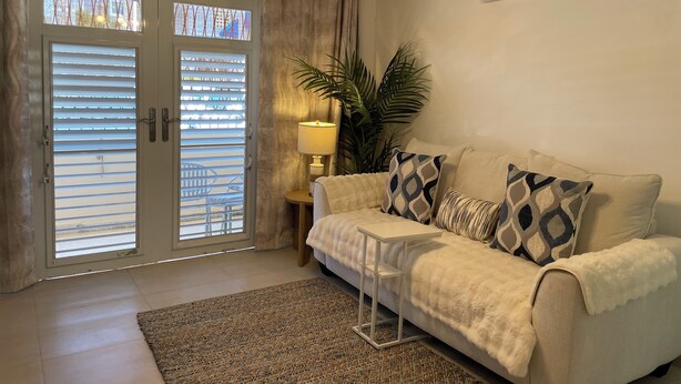 A living room with a beige sofa, patterned cushions, a small side table, a floor lamp, a ceiling fan, and a large window with blinds.