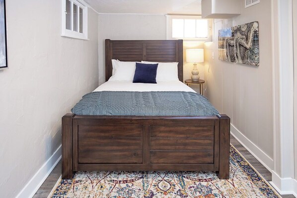 Une chambre à coucher avec un lit en bois, des murs blancs, un tapis à motifs et un tableau accroché au mur.