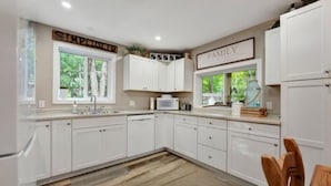 A modern kitchen with white cabinetry, a microwave, and a sink. There are two windows with views of greenery outside. - Peninsula Perch: Water & Trails (Wiarton)