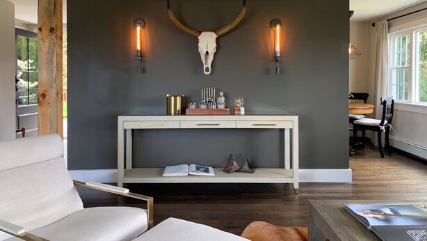 Un salon moderne agrémenté d’un grand crâne de cerf fixé au mur, de deux appliques murales, d’une console blanche ornée d’objets décoratifs et d’une grande fenêtre laissant entrer la lumière naturelle.