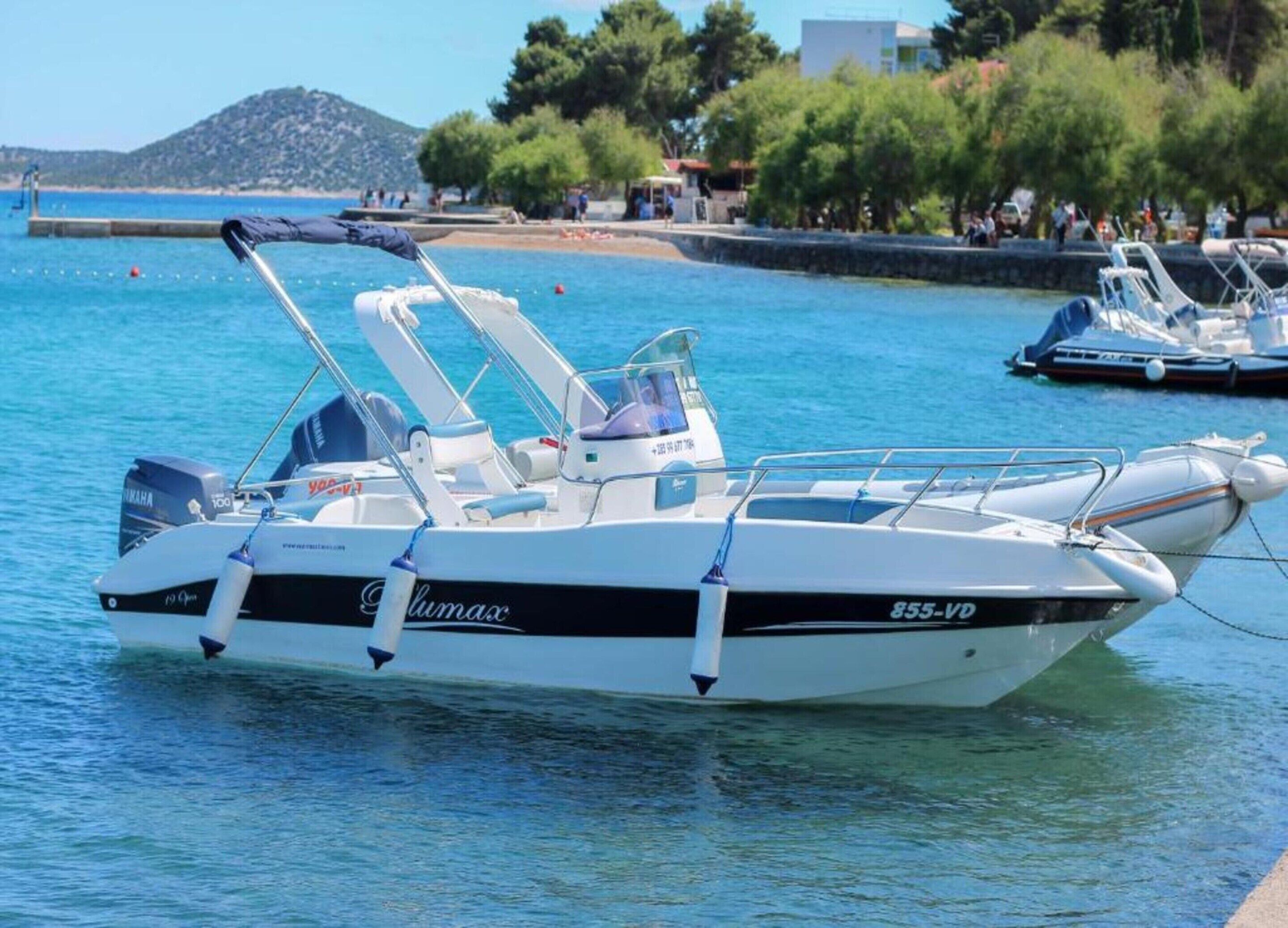 Ein Boot namens 'Gluma' liegt in einer Marina ab, im Hintergrund sind ruhiges Wasser, weitere Boote und ein Strand zu sehen.