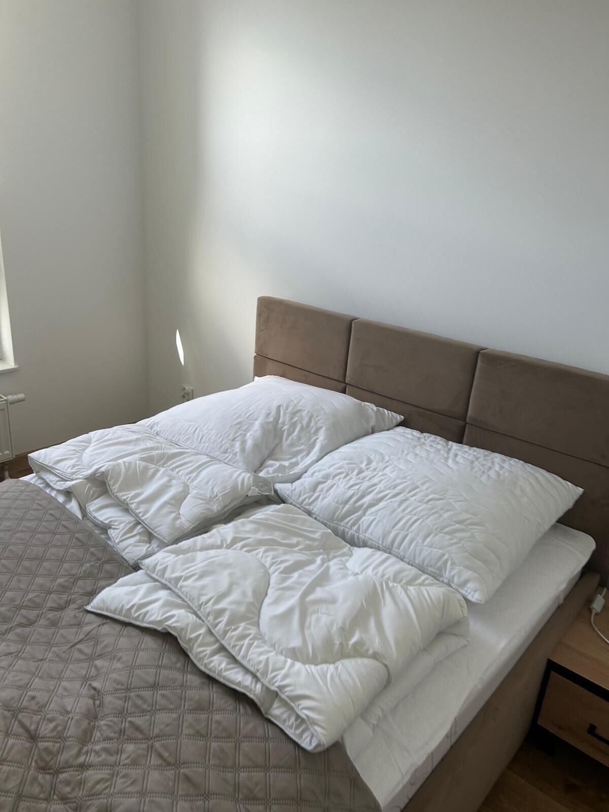 A neatly made bed with white bedding and a quilted blanket, a wooden nightstand with a drawer, and a plain wall with a window on the left.