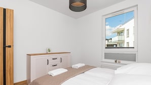 Une chambre à coucher comprenant un lit, une table de chevet, une commode, une fenêtre donnant sur des immeubles et un luminaire au plafond.
