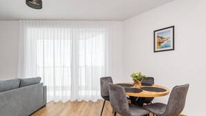 Un salon moderne avec un canapé gris, une table de salle à manger ronde en bois et quatre chaises grises, ainsi qu’une grande fenêtre ornée de rideaux blancs et légers.