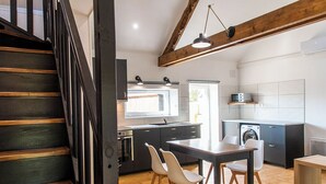A modern kitchen with dark cabinetry, a wooden staircase, and a dining area with white chairs. - Gîte on a farm with animals, near Arromanche-les-bains, 2 km from the sea (Ryes)
