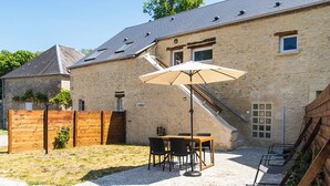 A stone building with a patio area featuring a table and chairs, an umbrella, and a wooden fence. - Gîte on a farm with animals, near Arromanche-les-bains, 2 km from the sea (Ryes)