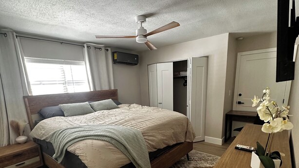 Une chambre à coucher comprenant un lit, des tables de chevet, une commode, un ventilateur de plafond, un appareil de climatisation, une fenêtre avec des rideaux, une porte et une petite table sur laquelle se trouvent une télécommande et une orchidée.