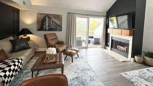 Un salon moderne comprenant un canapé, un fauteuil et un pouf, une cheminée et une grande fenêtre donnant sur l’extérieur.