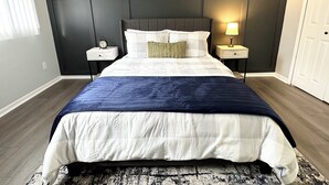Un lit bien fait, avec une couette bleue et blanche, deux tables de chevet avec des lampes, une horloge murale et un miroir décoratif.
