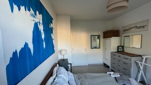 Un dormitorio con un cuadro abstracto grande, una cama con ropa de cama blanca, un aparador, un espejo y una lámpara colgante.
