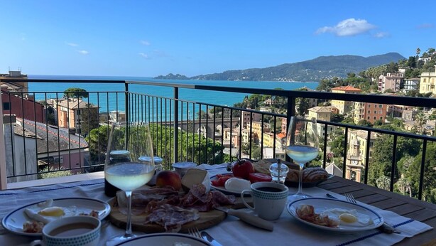 Una mesa en el balcón con alimentos para el desayuno, vista al mar y edificios al fondo.