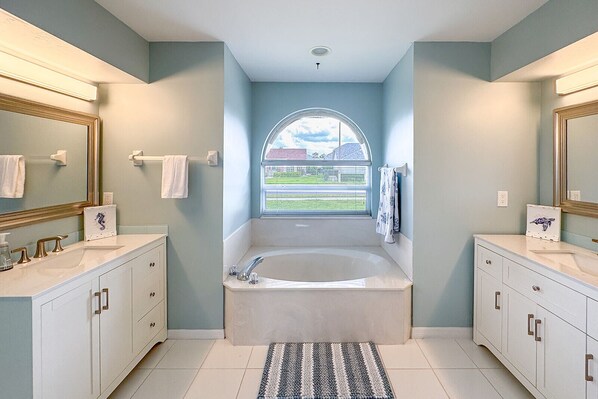 A modern bathroom with a large bathtub, two sinks, and a window with a view of a building and greenery.