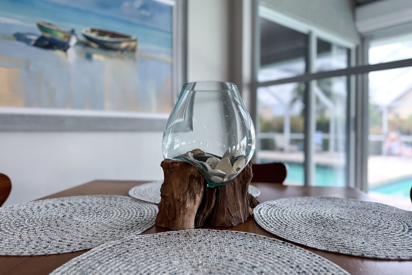 A glass sculpture on a wooden stand, surrounded by woven placemats on a table with a view of a swimming pool through large windows.