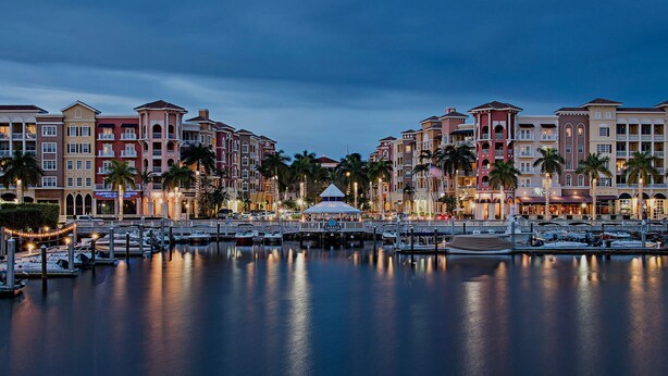 A marina with boats docked, a gazebo, and multi-story buildings with balconies and palm trees.