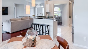 A modern kitchen with white cabinets, a central island, and bar stools. There is a dining table with a glass vase and a woven placemat.