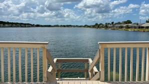 A waterfront view from a wooden deck with a railing, overlooking a calm lake and a row of houses with a clear sky above.