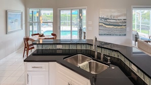 A modern kitchen with a black countertop, stainless steel sink, and white cabinetry. There is a dining area with a table and chairs visible through large windows.