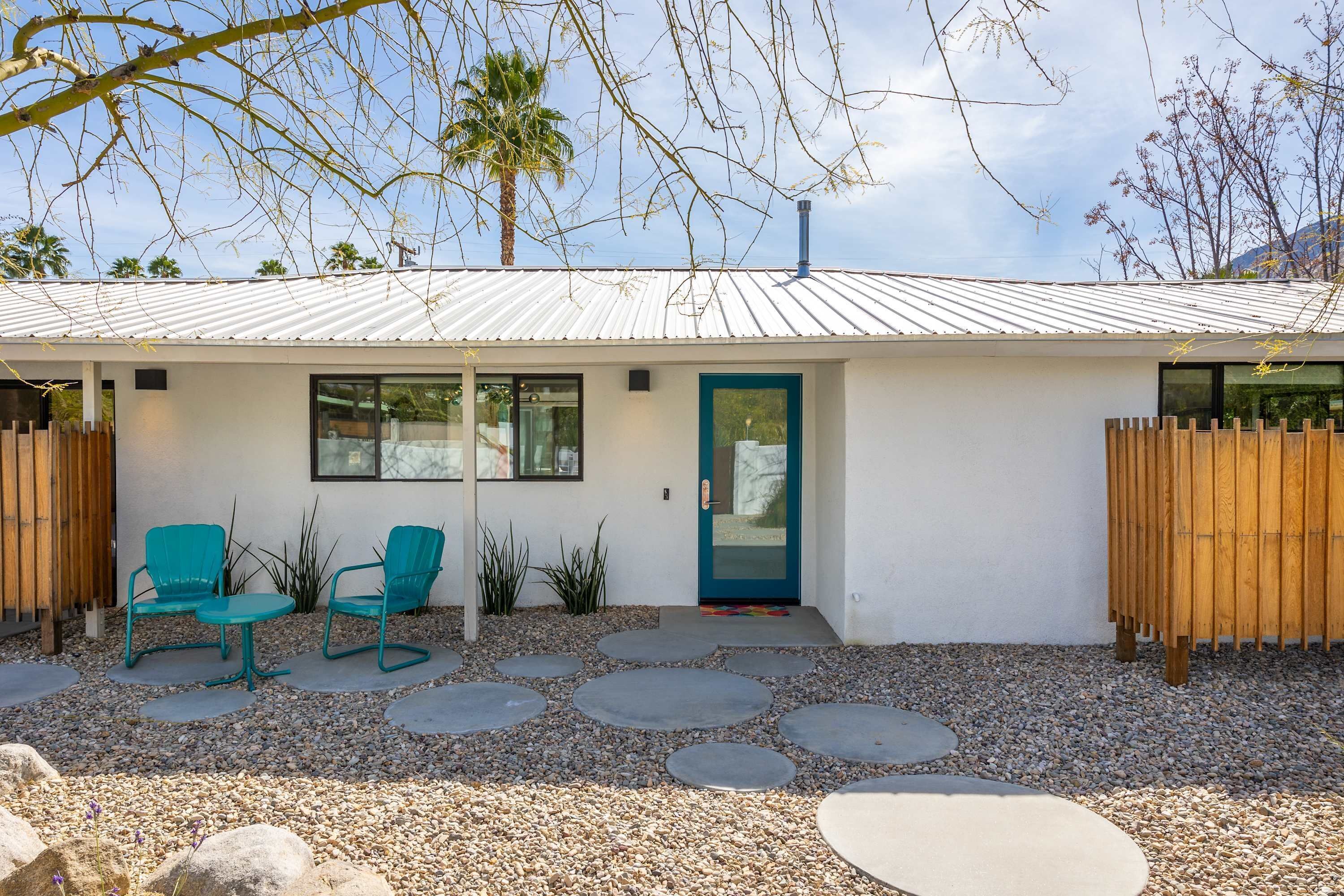 A modern house with a white exterior, a blue door, and a small patio area with two green chairs and a table.