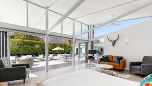 A modern living room with a large bed, two sofas, a coffee table, and a TV. There is a glass wall with a view of a pool and greenery. - Luxury 4-bedroom Home With Pool Spa Stunning Mountain Views (Palm Springs)