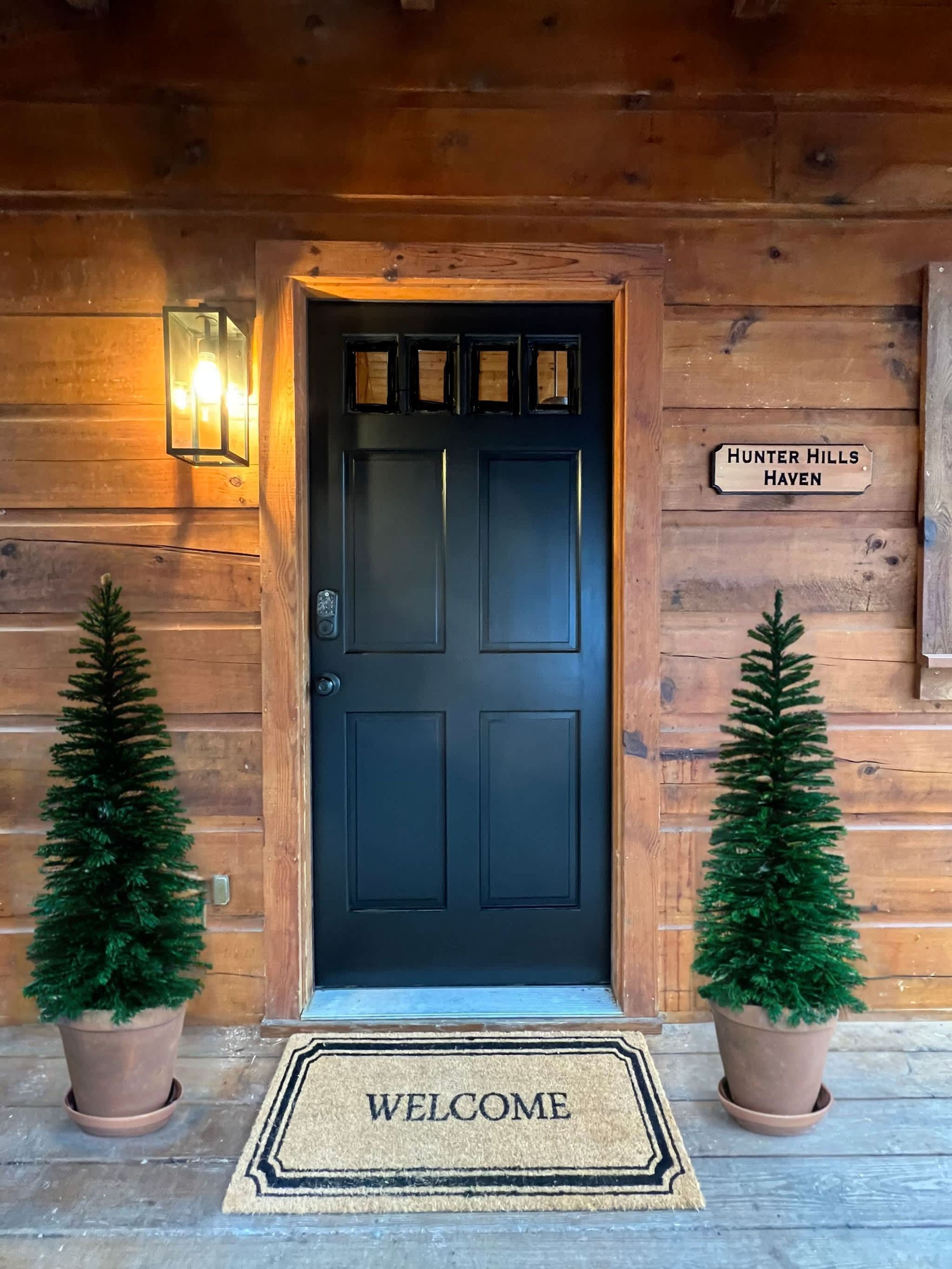 Entrada a una cabaña de madera con una puerta negra, un felpudo y un cartel que dice "Hunter Hills Haven".