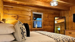 A wooden cabin bedroom with a bed, pillows, a window, and a mirror. - Hunter Hills Haven - Secluded Luxe Cabin Getaway With Mountain Views Spa Hot Tub Game Room (Sevierville)