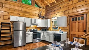 A wooden cabin-style kitchen with white cabinets, stainless steel appliances, and a dining area with a table set for four. - Hunter Hills Haven - Secluded Luxe Cabin Getaway With Mountain Views Spa Hot Tub Game Room (Sevierville)