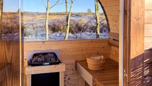 Una sauna de madera con vistas a un paisaje natural, un calentador negro y un cubo de madera.