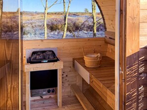 Una sauna de madera con vistas a un paisaje natural, un calentador negro y un cubo de madera.