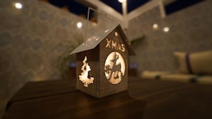 A wooden house-shaped decoration with 'XMAS' and a deer silhouette, placed on a table in a dimly lit room. - Anza Sets House  (Anza)