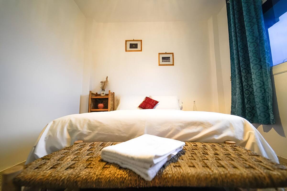 A neatly made bed with white linens, a small bedside table with a vase and a red pillow, and two framed pictures on the wall.