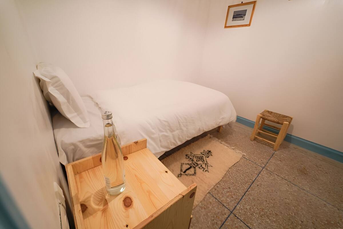 A bedroom with a bed, a wooden nightstand, a small stool, and a framed picture on the wall.