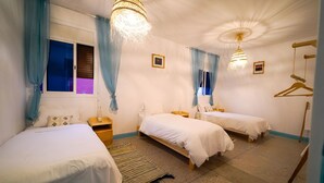 A hotel room with two single beds, a wooden nightstand, a hanging light fixture, and a window with blue curtains. - Anza Sets House  (Anza)