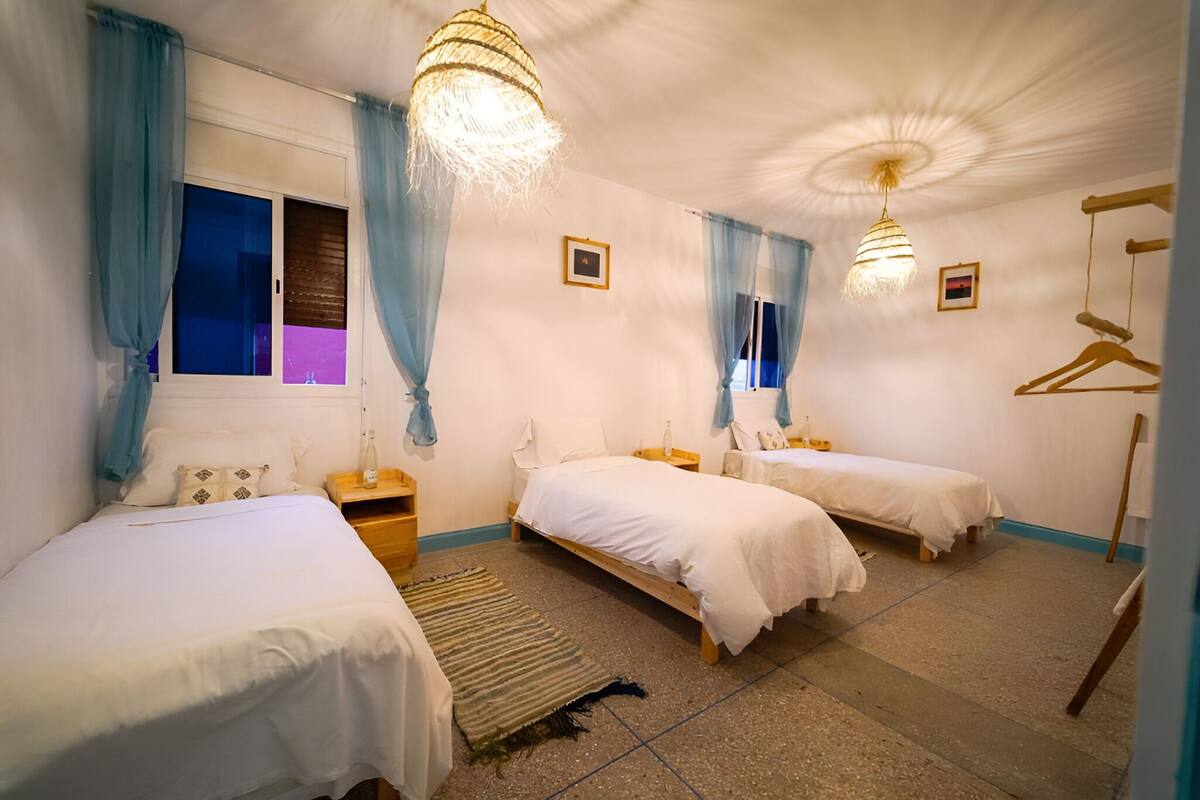 A hotel room with two single beds, a wooden nightstand, a hanging light fixture, and a window with blue curtains.