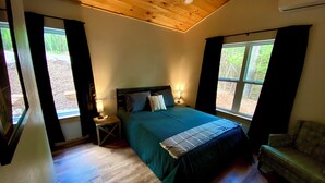 A bedroom with a wooden ceiling, a bed with a blue bedspread, two bedside lamps, a nightstand, a window with a view of trees, and a green armchair. - Cedar Hill Cabin 2 bed 2 bath cabin in the Ozarks • Hot Tub • (Harrison)