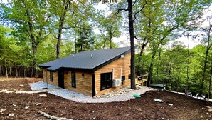 A small wooden cabin with a black roof, surrounded by trees and a gravel path. - Cedar Hill Cabin 2 bed 2 bath cabin in the Ozarks • Hot Tub • (Harrison)