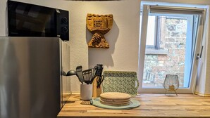 A kitchen with a refrigerator, a microwave, a wooden cutting board, a knife block, and a window with a view of a stone building.