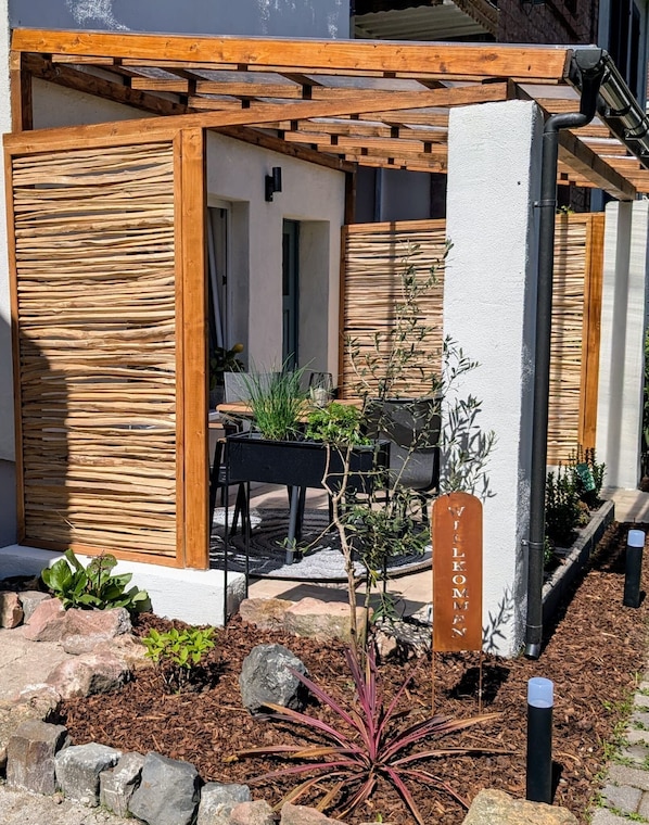 A covered outdoor seating area with a pergola, a welcome sign, and a garden with plants and rocks.