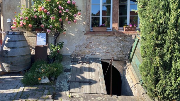 A rustic building with a stone wall, a wooden barrel, and a small wooden bridge leading to a door.