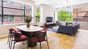 A spacious living area with a round dining table, red chairs, a sofa, and a TV. Large windows offer views of a brick building and greenery outside.