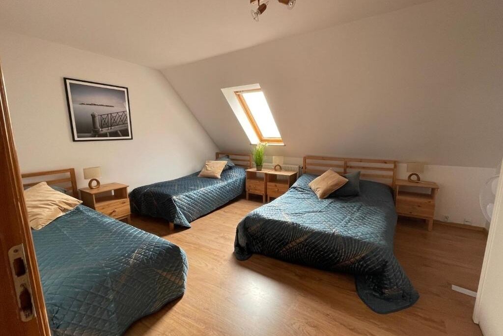 Et værelse med tre senge, trægavle, et ovenlys, et indrammet billede på væggen og et lille bord med en lampe.