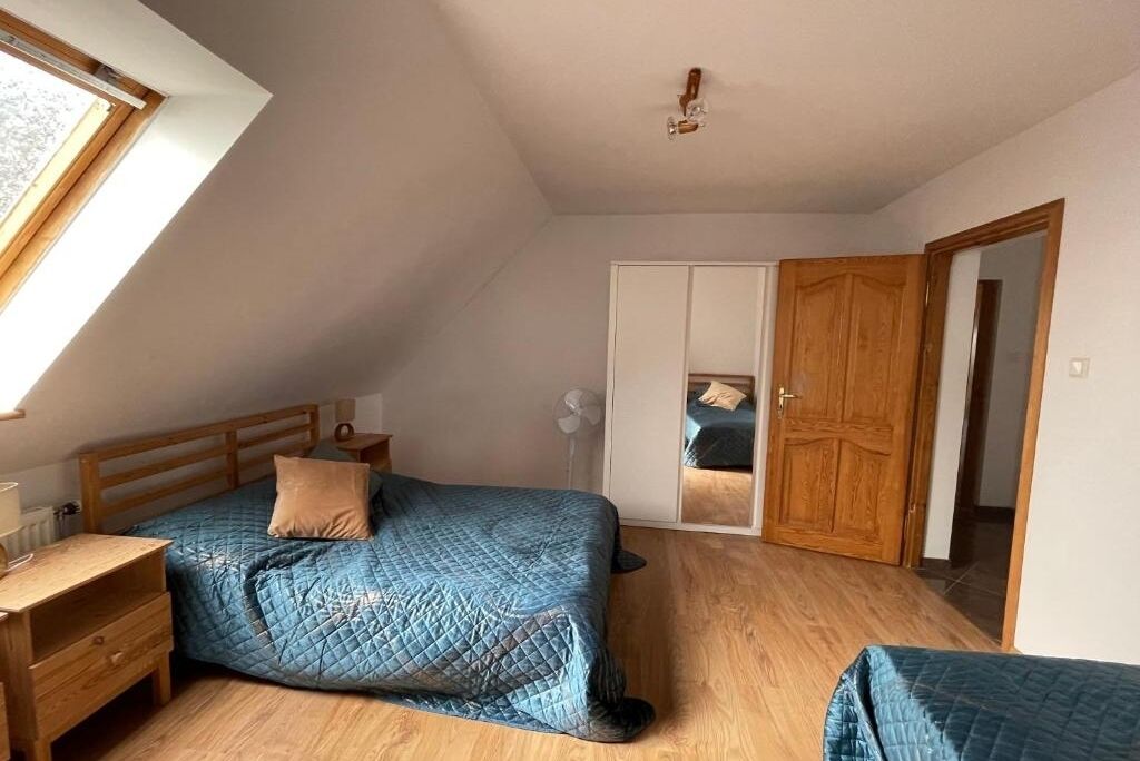 A bedroom with a sloped ceiling, a bed with a blue quilt, a wooden nightstand, a wardrobe, and a door leading to another room.