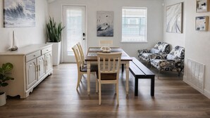 A dining area with a wooden table and chairs, a bench, a sofa, and a cabinet. There are two windows with blinds, a door, and a plant.