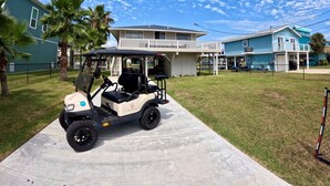 A golf cart parked on a concrete path in front of a house with a balcony, surrounded by palm trees and other houses.