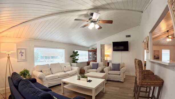 A spacious living room with a high ceiling, a ceiling fan, and a TV mounted on the wall. There are multiple seating options including sofas and chairs, and a coffee table in the center.